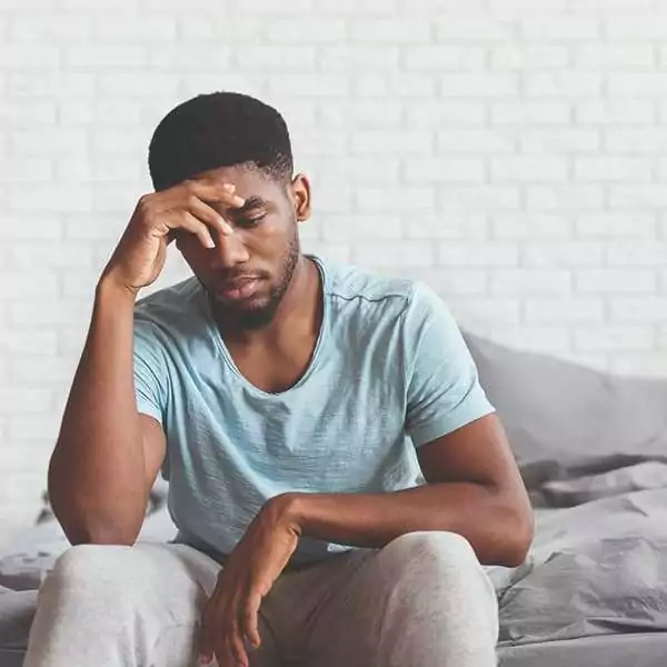 Erectile Dysfunction, young black male sitting on the bottom of his bed with his left hand across his face looking sad